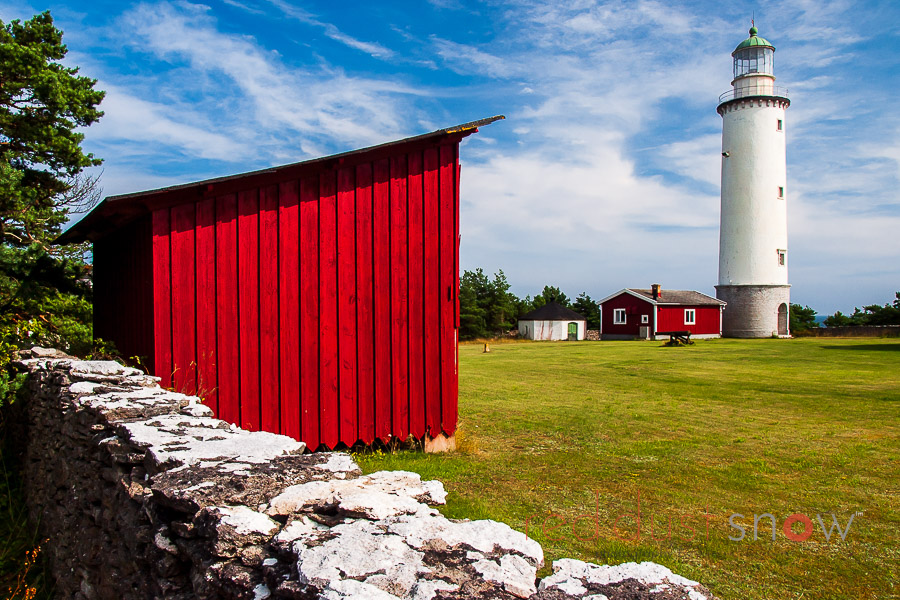 Red painted shed (Stuga) in the foreground with a lighthouse in the distance. Gotland, Sweden.